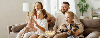 Deux adultes et deux enfants son assis dans un capé et regardent la télé en mangeant des pop-corn.