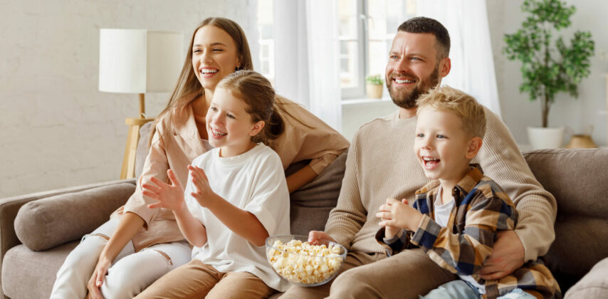 Deux adultes et deux enfants son assis dans un capé et regardent la télé en mangeant des pop-corn.